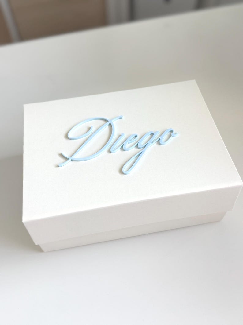 May include: A white gift box with a light blue cursive "Diego" on the lid. The box is rectangular and sits on a white surface. The text is in a delicate, flowing script.