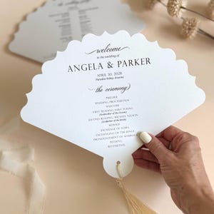 May include: White, fan-shaped wedding program with scalloped edges. The program includes the names "Angela & Parker", the date, and ceremony details. A tassel hangs from the bottom. The program is held by a person.