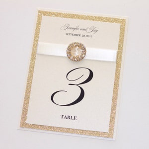 May include: White wedding table number card with gold glitter border. The card has a black number "3" and the word "TABLE" printed on it. A white satin ribbon with a rhinestone-encrusted button is attached to the top of the card. The card says "Jennifer and Jay" and "SEPTEMBER 28, 2013" at the top.