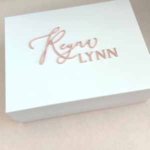 May include: A white gift box with a rose gold name inscription that reads "Reyna Lynn". The box is rectangular with a slightly raised lid. The inscription is in a cursive font for the name "Reyna" and a block font for "Lynn".