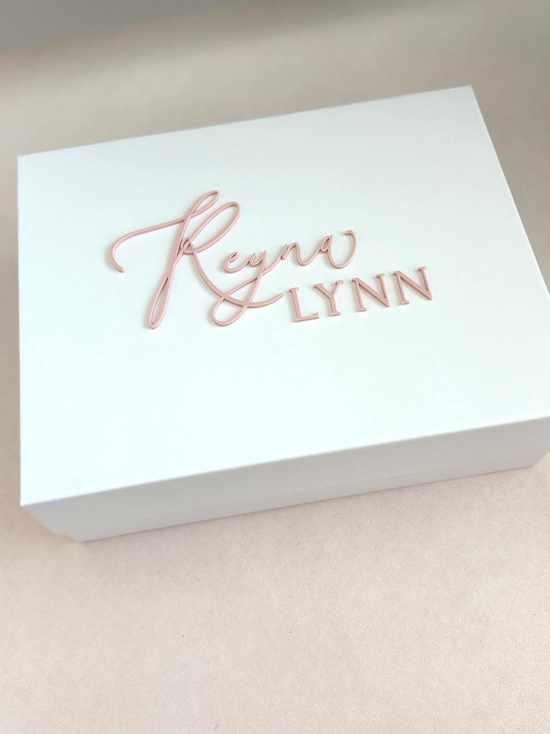 May include: A white gift box with a rose gold name inscription that reads "Reyna Lynn". The box is rectangular with a slightly raised lid. The inscription is in a cursive font for the name "Reyna" and a block font for "Lynn".