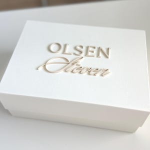 May include: A white box with the name "OLSEN Steven" embossed on the lid in a light beige color. The box is rectangular and appears to be made of a sturdy material. The overall aesthetic is clean and minimalist.