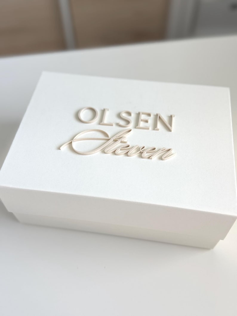 May include: A white box with the name "OLSEN Steven" embossed on the lid in a light beige color. The box is rectangular and appears to be made of a sturdy material. The overall aesthetic is clean and minimalist.