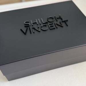 May include: A black box with the name "SHILOH VINCENT" embossed on the lid. The box is rectangular and appears to be made of a matte material. The text is in a sans-serif font.