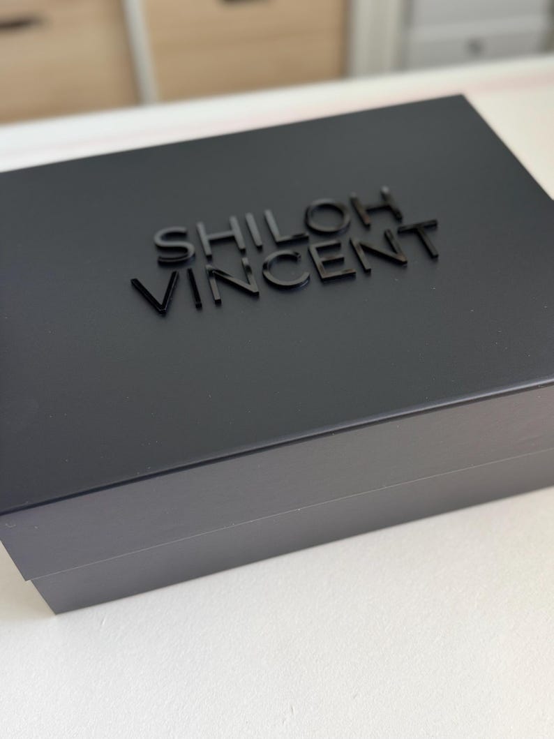 May include: A black box with the name "SHILOH VINCENT" embossed on the lid. The box is rectangular and appears to be made of a matte material. The text is in a sans-serif font.