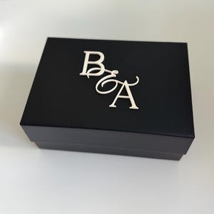 May include: A black box with the letters "B" and "A" in a cursive font on the top. The letters are in a silver color. The box is sitting on a white surface.