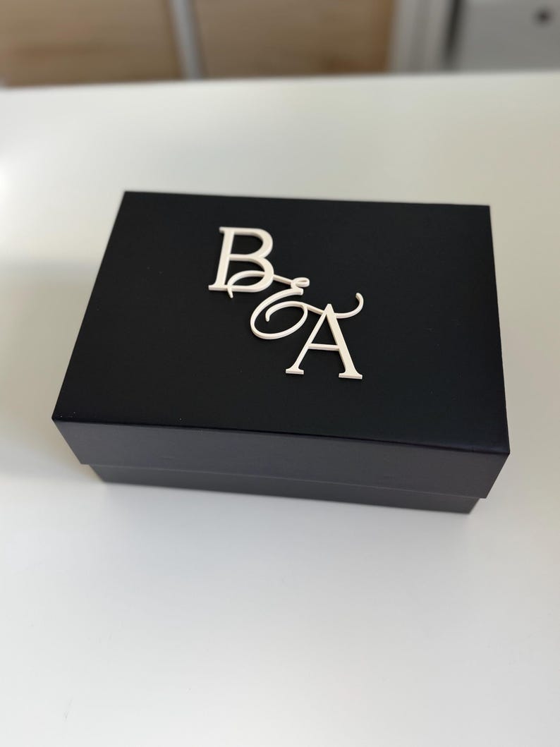May include: A black box with the letters "B" and "A" in a cursive font on the top. The letters are in a silver color. The box is sitting on a white surface.