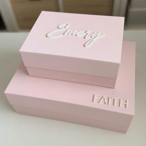 May include: Two stacked, light pink boxes with white lettering. The top box reads "Emery" in cursive, and the bottom box reads "FAITH" in block letters. The boxes are on a white surface.