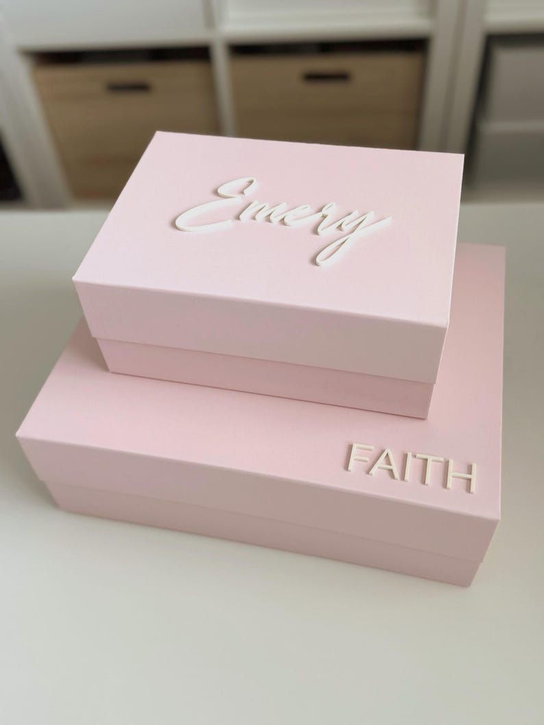 May include: Two stacked, light pink boxes with white lettering. The top box reads "Emery" in cursive, and the bottom box reads "FAITH" in block letters. The boxes are on a white surface.