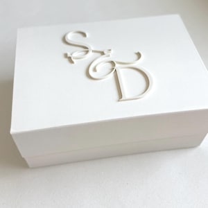 May include: A white square box with the embossed letters "S" and "D" in a cursive font on the lid. The box is plain and minimalist, with clean lines and a simple design. The box is likely for jewelry or small gifts.