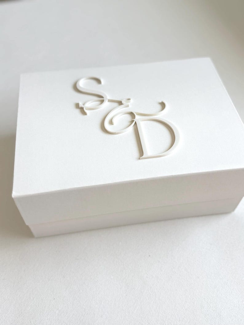 May include: A white square box with the embossed letters "S" and "D" in a cursive font on the lid. The box is plain and minimalist, with clean lines and a simple design. The box is likely for jewelry or small gifts.