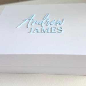 May include: A white gift box with the name "Andrew JAMES" in light blue script lettering on the top. The box is rectangular and has a clean, minimalist design, suitable for various occasions.