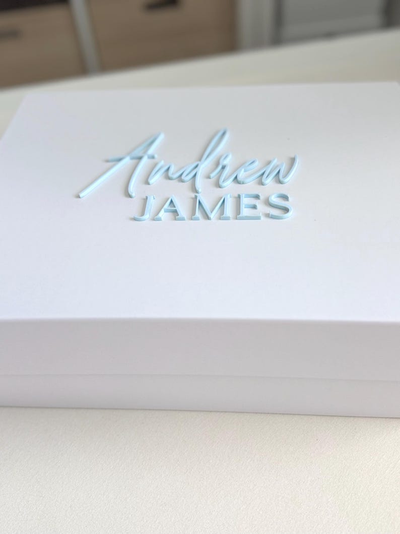 May include: A white gift box with the name "Andrew JAMES" in light blue script lettering on the top. The box is rectangular and has a clean, minimalist design, suitable for various occasions.