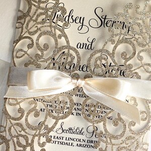 May include: Laser cut wedding invitation with gold glitter and a white lace design. The invitation features the names Lindsey Sterling and Michael Stone, the wedding date, and the location of the wedding. The text "Reply" is on a separate card with a space for the guest to RSVP.