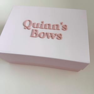 May include: A light pink box with the words "Quinn's Bows" in raised, matching pink lettering. The box is rectangular with a lid and sits on a white surface. The overall aesthetic is soft and feminine.