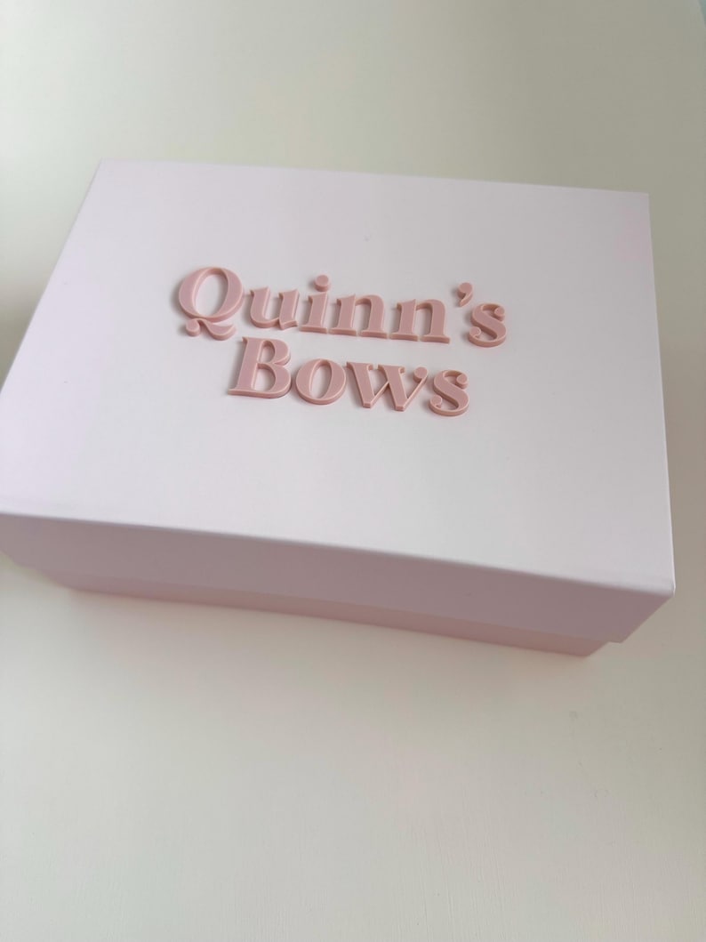 May include: A light pink box with the words "Quinn's Bows" in raised, matching pink lettering. The box is rectangular with a lid and sits on a white surface. The overall aesthetic is soft and feminine.