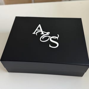 May include: A black rectangular box with a white monogram of the letters "AoS" on the top. The box is closed and sits on a white surface. The background is blurred, showing a glimpse of a light-colored wall and cabinets.
