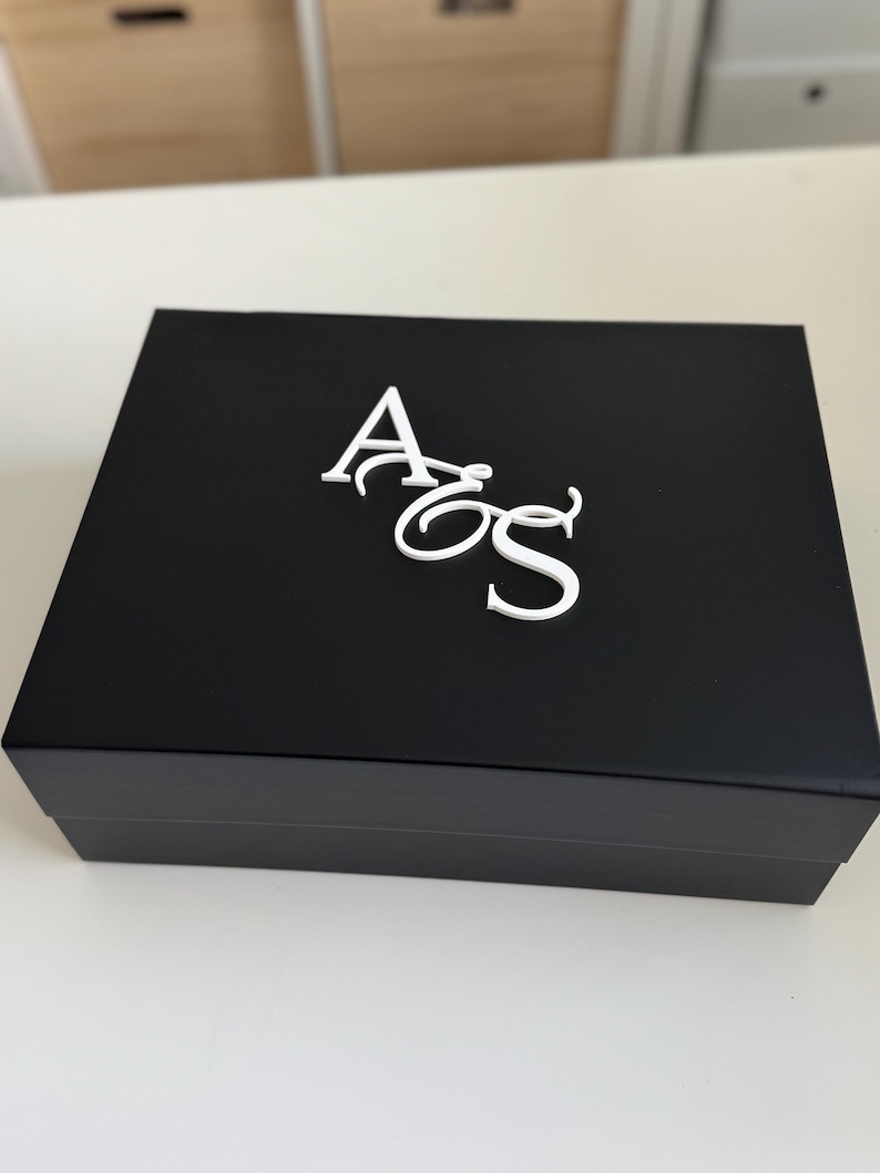 May include: A black rectangular box with a white monogram of the letters "AoS" on the top. The box is closed and sits on a white surface. The background is blurred, showing a glimpse of a light-colored wall and cabinets.