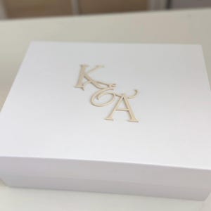May include: A white gift box with gold-colored initials 'K' and 'A' on the lid. The box is rectangular and appears to be made of a sturdy material. The initials are in a decorative font.