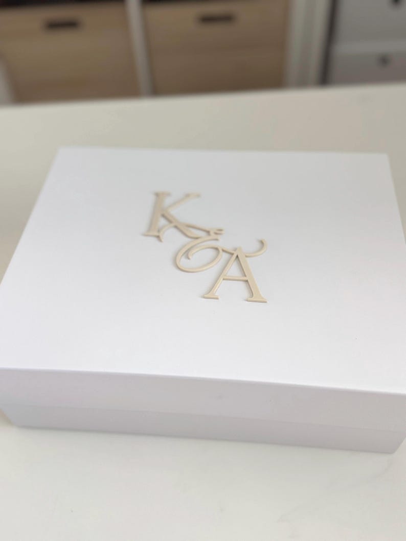 May include: A white gift box with gold-colored initials 'K' and 'A' on the lid. The box is rectangular and appears to be made of a sturdy material. The initials are in a decorative font.