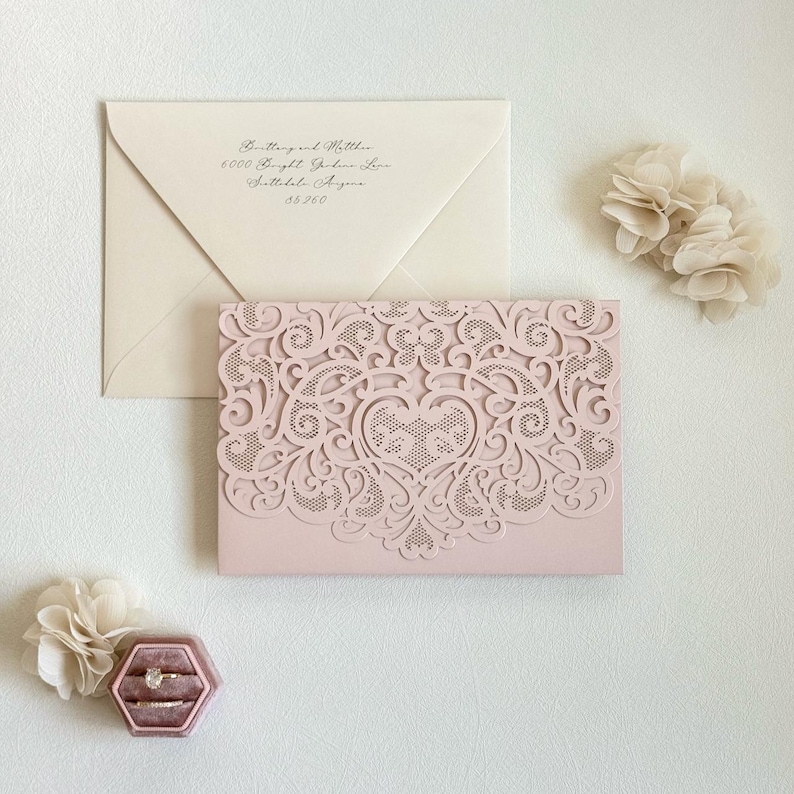 May include: A blush pink wedding invitation with a delicate laser-cut design featuring a heart and intricate floral patterns. The invitation is paired with a white envelope.