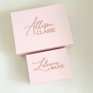 May include: Two pink gift boxes, one larger and one smaller, are displayed. The larger box has the name "Allison CLAIRE" in a cursive font. The smaller box has the name "Liliana MARIE" in a similar style. The boxes are on a white surface.