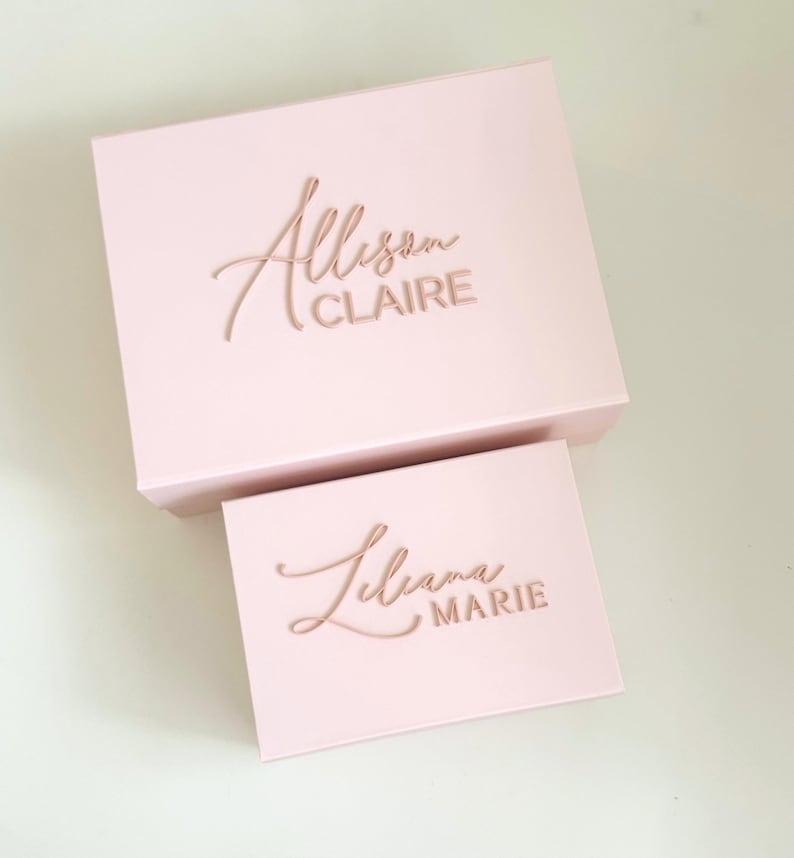 May include: Two pink gift boxes, one larger and one smaller, are displayed. The larger box has the name "Allison CLAIRE" in a cursive font. The smaller box has the name "Liliana MARIE" in a similar style. The boxes are on a white surface.