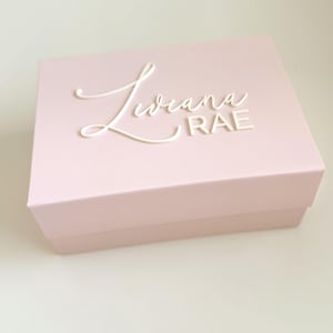 May include: A light pink rectangular box with a lid. The box has the name "Liviana RAE" in white cursive lettering on the top. The box is sitting on a white surface.