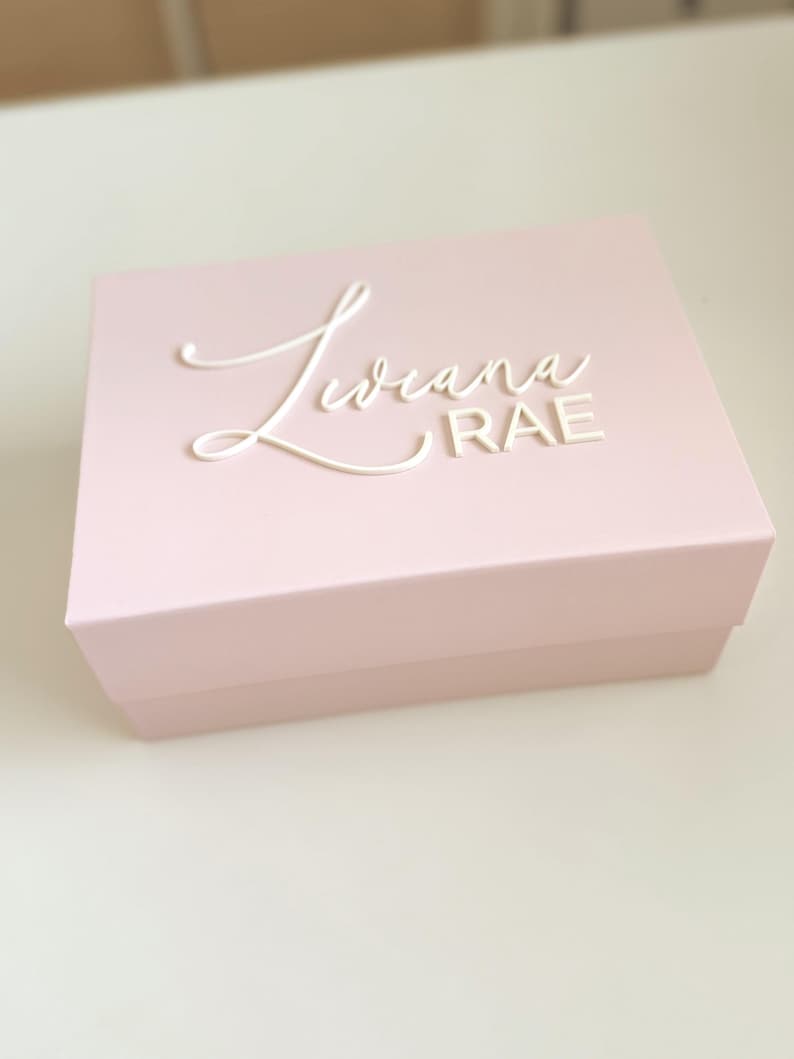 May include: A light pink rectangular box with a lid. The box has the name "Liviana RAE" in white cursive lettering on the top. The box is sitting on a white surface.