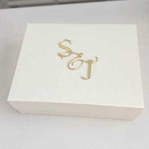 May include: A cream-colored box with a gold monogram of the letters "S" and "T" on the lid. The box is rectangular and appears to be made of cardboard or a similar material. The gold monogram is in a cursive font.