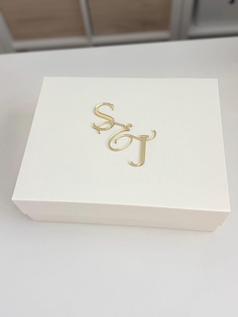 May include: A cream-colored box with a gold monogram of the letters "S" and "T" on the lid. The box is rectangular and appears to be made of cardboard or a similar material. The gold monogram is in a cursive font.