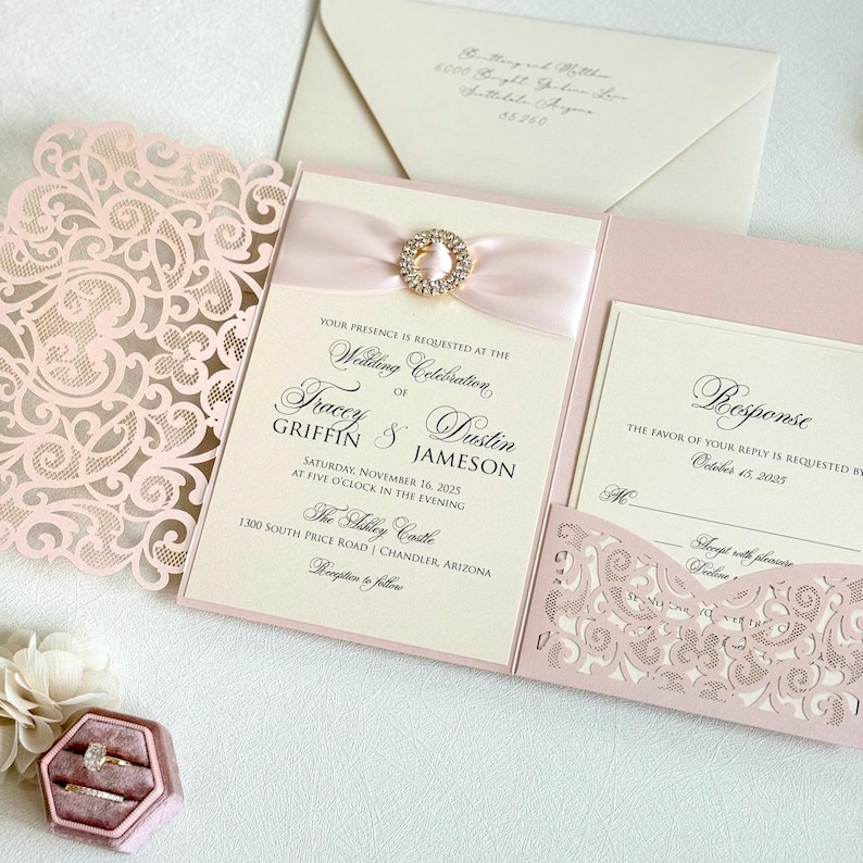 May include: Pink and ivory wedding invitation with a laser cut design. The invitation features a pink ribbon with a rhinestone embellishment. The text on the invitation reads "Wedding Celebration of Tracey & Dustin Griffin Jameson, Saturday, November 16, 2025 at five o'clock in the evening, The Abbey Castle, 1300 South Price Road, Chandler, Arizona, Reception to follow". The RSVP card reads "Response, The favor of your reply is requested by October 15, 2025".
