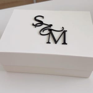 May include: A white rectangular gift box with a black monogram of the letters "S" and "M" on the lid. The box is plain and simple, with a clean, modern aesthetic. The letters are in a cursive font.