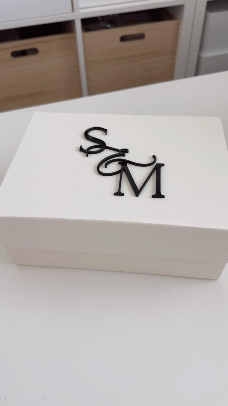 May include: A white rectangular gift box with a black monogram of the letters "S" and "M" on the lid. The box is plain and simple, with a clean, modern aesthetic. The letters are in a cursive font.