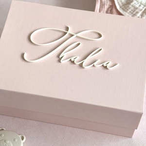 May include: A light pink baby keepsake box with the name "Thalia Rose" in white script. The box is next to a white crochet bunny and a bear-shaped teether. The image also includes text that says "Baby Keepsake Box" and "Baby Shower Gift Idea".