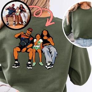May include: Olive green sweatshirt featuring a cartoon illustration of a family. The illustration depicts a man, a woman, and a child sitting together. A smaller image of the family is in a circle above the illustration.