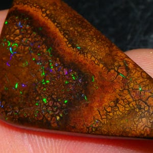 Puede incluir: Una piedra preciosa de ópalo boulder triangular que presenta una matriz de piedra de hierro marrón oscuro con una red compleja de grietas. La superficie muestra destellos vibrantes de colores verde y morado iridiscentes en toda la piedra.