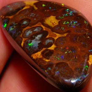 Könnte beinhalten: Ein polierter, tropfenförmiger Opal mit einer satten braunen Grundfarbe. Der Stein weist ein einzigartiges Muster aus goldgelben und dunkelbraunen Wirbeln mit grünen, blauen und roten irisierenden Blitzen auf. Der Opal wird in einer Hand gehalten.