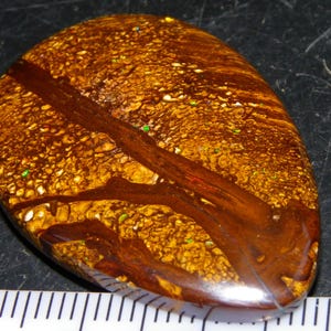 May include: A polished, teardrop-shaped boulder opal gemstone featuring a rich brown ironstone matrix with swirling dark brown veins and scattered flecks of iridescent green and gold color, positioned above a ruler showing inches.
