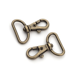 May include: Two antique gold-toned metal swivel clasps. The clasps are open and ready to be attached to a bag, strap, or other item.