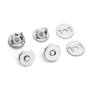 May include: A collection of silver-colored magnetic snap closures for bags or other craft projects. The set includes several round pieces, some with prongs and others with a central hole for sewing or attachment. The metal has a shiny finish.