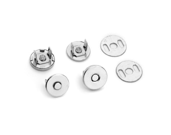 May include: A collection of silver-colored magnetic snap closures for bags or other craft projects. The set includes several round pieces, some with prongs and others with a central hole for sewing or attachment. The metal has a shiny finish.