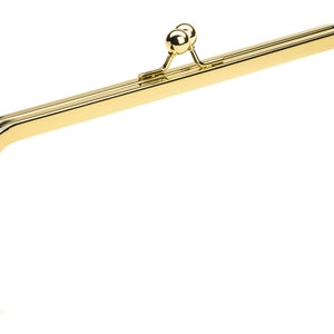 May include: A shiny, gold-colored purse frame with a classic design. The frame has a rectangular shape with rounded corners and a decorative clasp with two small, round balls. The frame is made of metal and has a smooth, polished finish.
