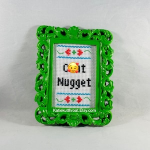 May include: A green framed cross-stitch with the text "Cool It Nugget" and a smiling emoji. The cross-stitch is framed in a green ornate frame.