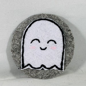 May include: A white felt ghost with a black embroidered smiling face and rosy cheeks, attached to a gray felt circle.
