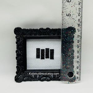 May include: A small black ornate frame containing a white cross-stitch fabric with four black vertical bars. A clear ruler with colourful glitter is placed next to the frame, showing the height is approximately 6.5 cm tall.