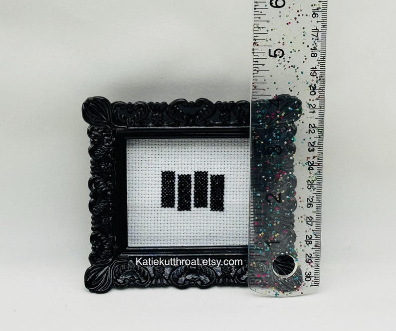 May include: A small black ornate frame containing a white cross-stitch fabric with four black vertical bars. A clear ruler with colourful glitter is placed next to the frame, showing the height is approximately 6.5 cm tall.