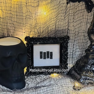 May include: A matte black skull-shaped candle holder, a small square framed cross-stitch featuring four vertical black bars on a white grid, and a black ornate lamp base. The text Katiekutthroat.etsy.com is visible on the frame.