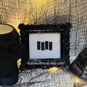 May include: A black ornate frame containing a white cross-stitch fabric piece with four vertical black bars embroidered in the centre. The text Katiekutthroat.etsy.com is printed in white at the bottom of the frame's inner border area.