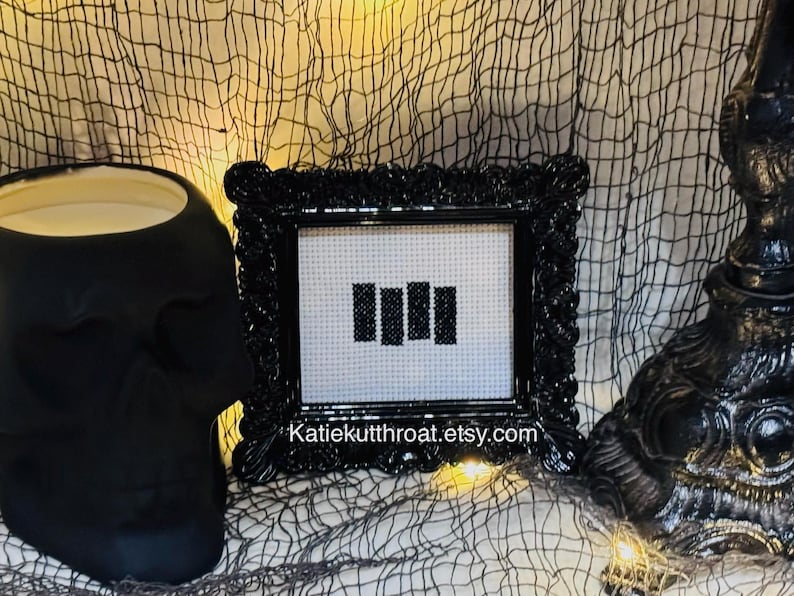 May include: A black ornate frame containing a white cross-stitch fabric piece with four vertical black bars embroidered in the centre. The text Katiekutthroat.etsy.com is printed in white at the bottom of the frame's inner border area.